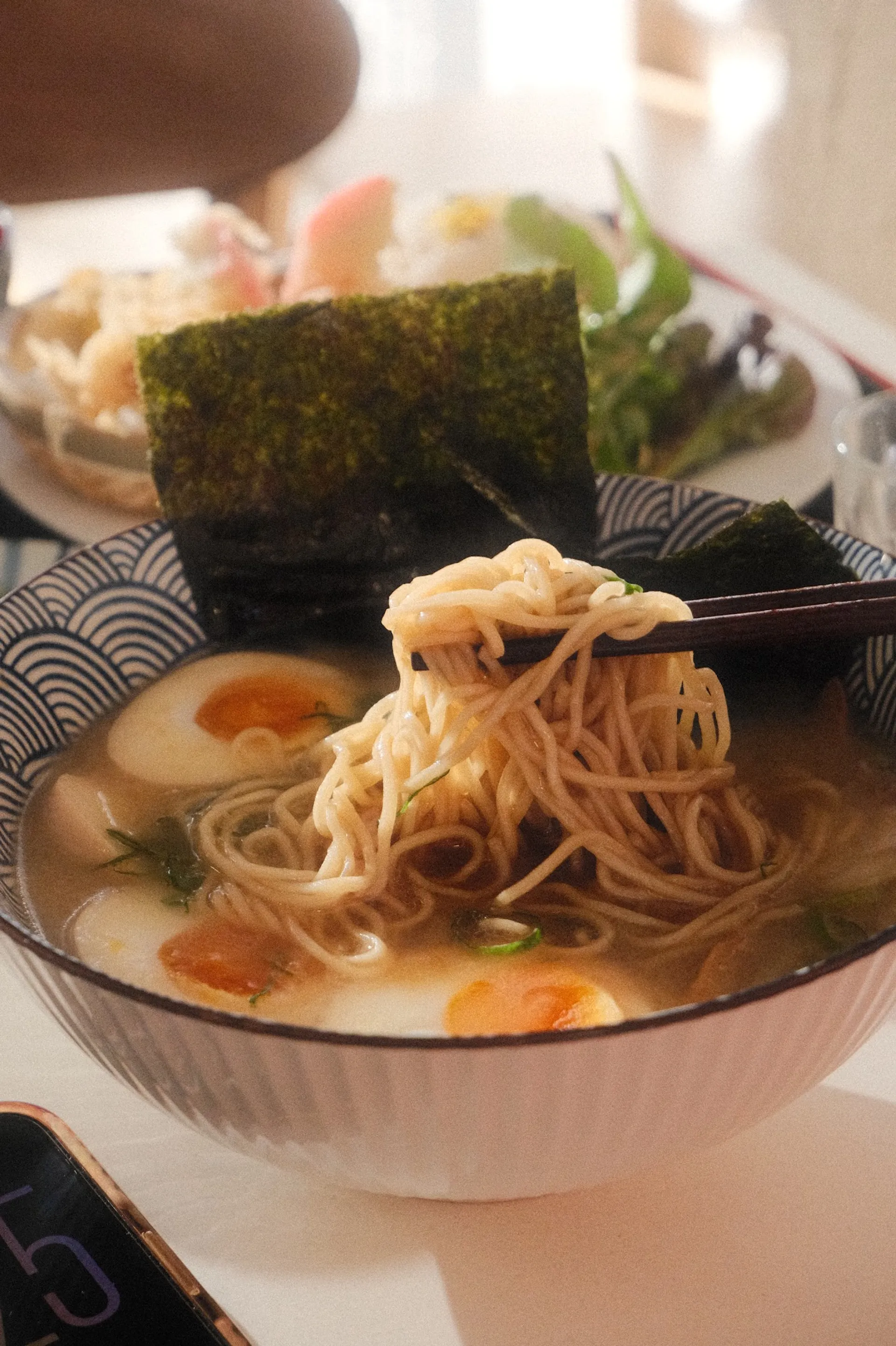 Ramen noodles lifted by chopsticks above broth with egg and nori
